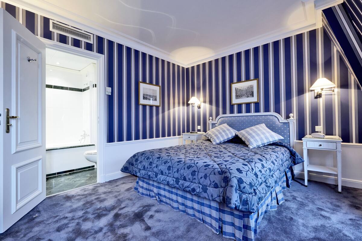 A bedroom with a bed, bedside tables, a nightstand, a lamp, and a door leading to a bathroom.