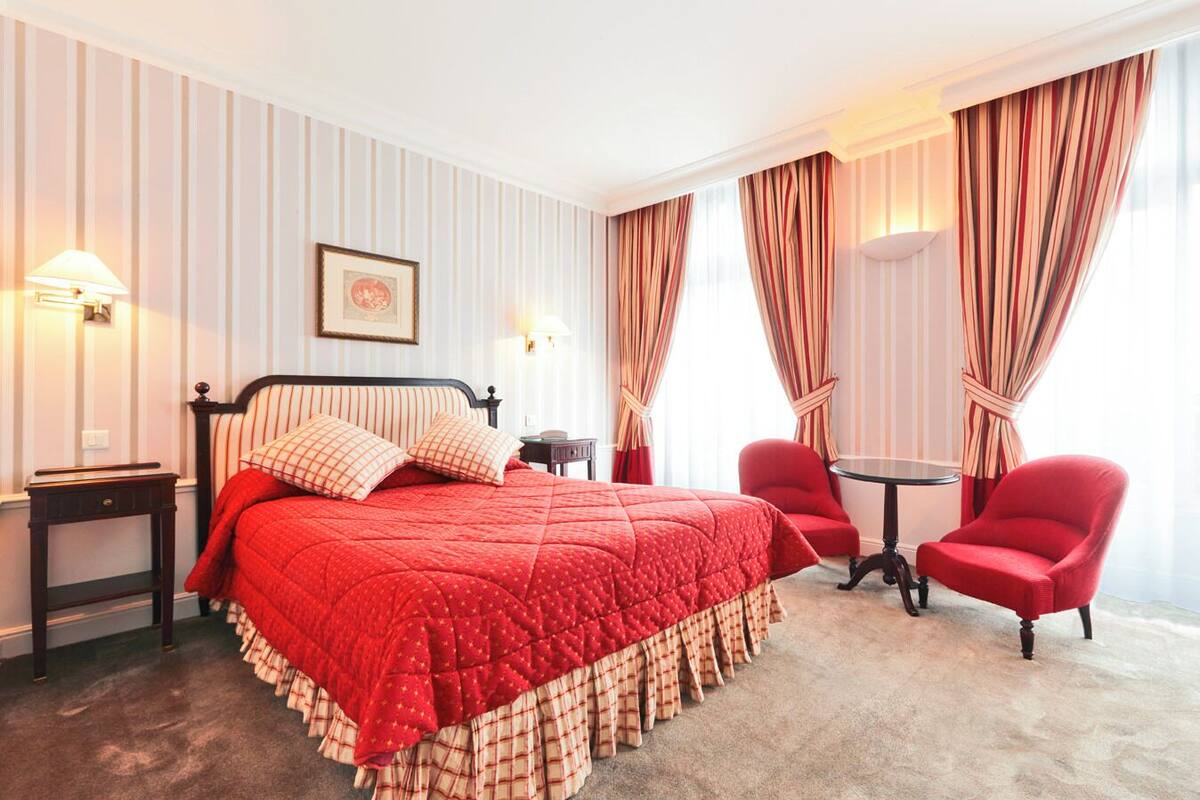 A hotel room with a large bed, two red chairs, a small table, and striped curtains.