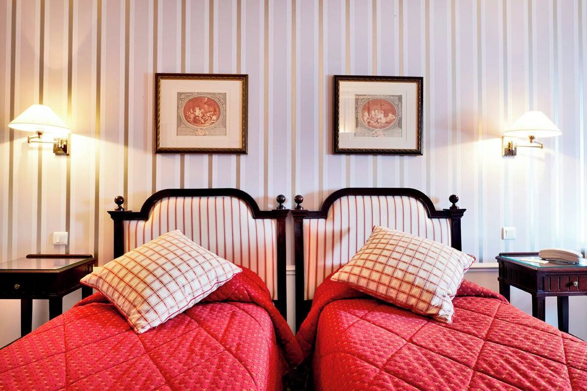 A double bed with red bedding and checkered pillows, flanked by two nightstands with lamps, against a striped wallpaper background.