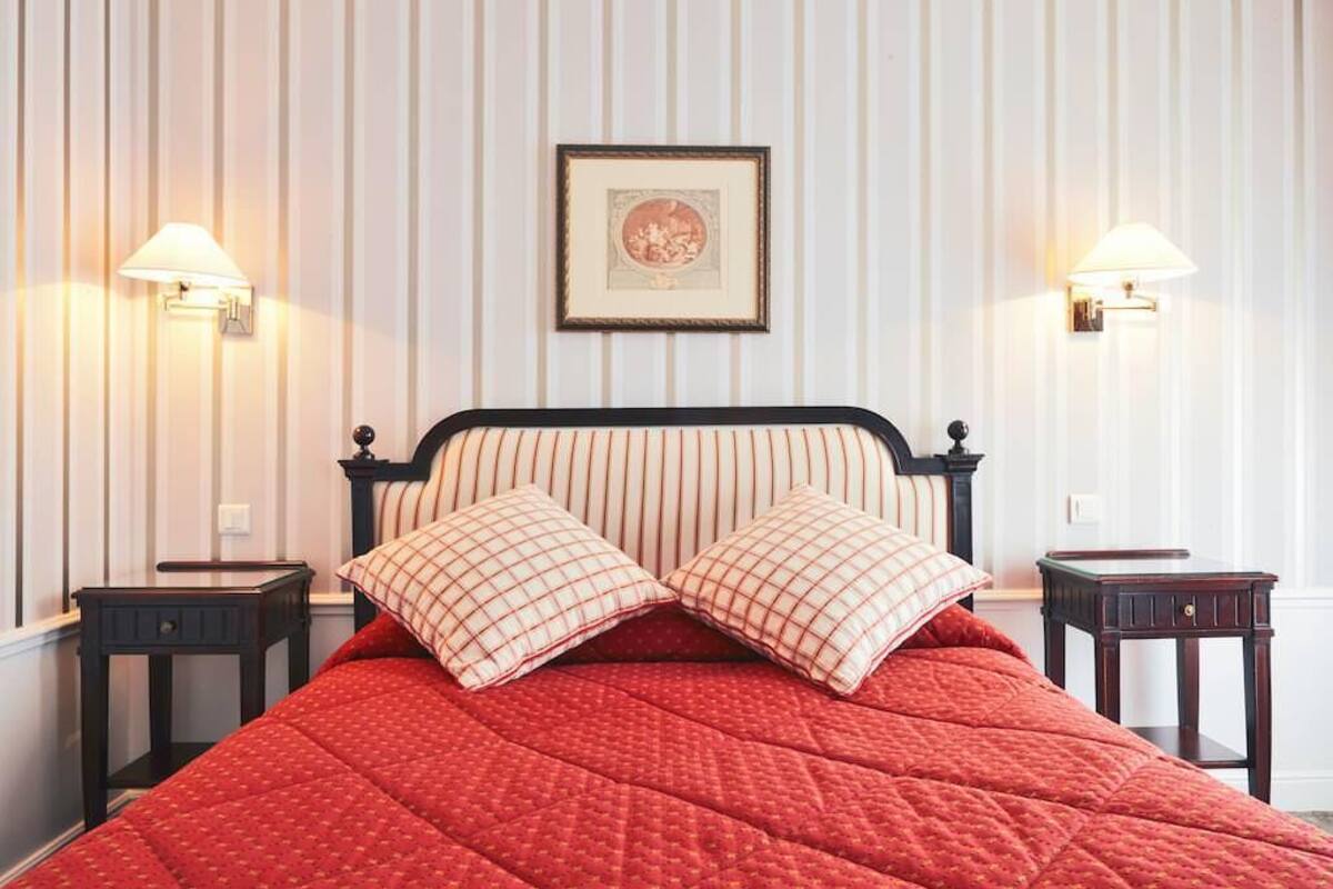 A neatly made bed with a red quilt, two matching pillows, and a framed picture above it. There are two wall-mounted lamps on either side of the bed.