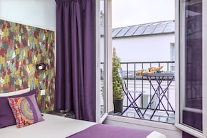A bedroom with a large window, a balcony with a table and chairs, and a colorful floral wallpaper.