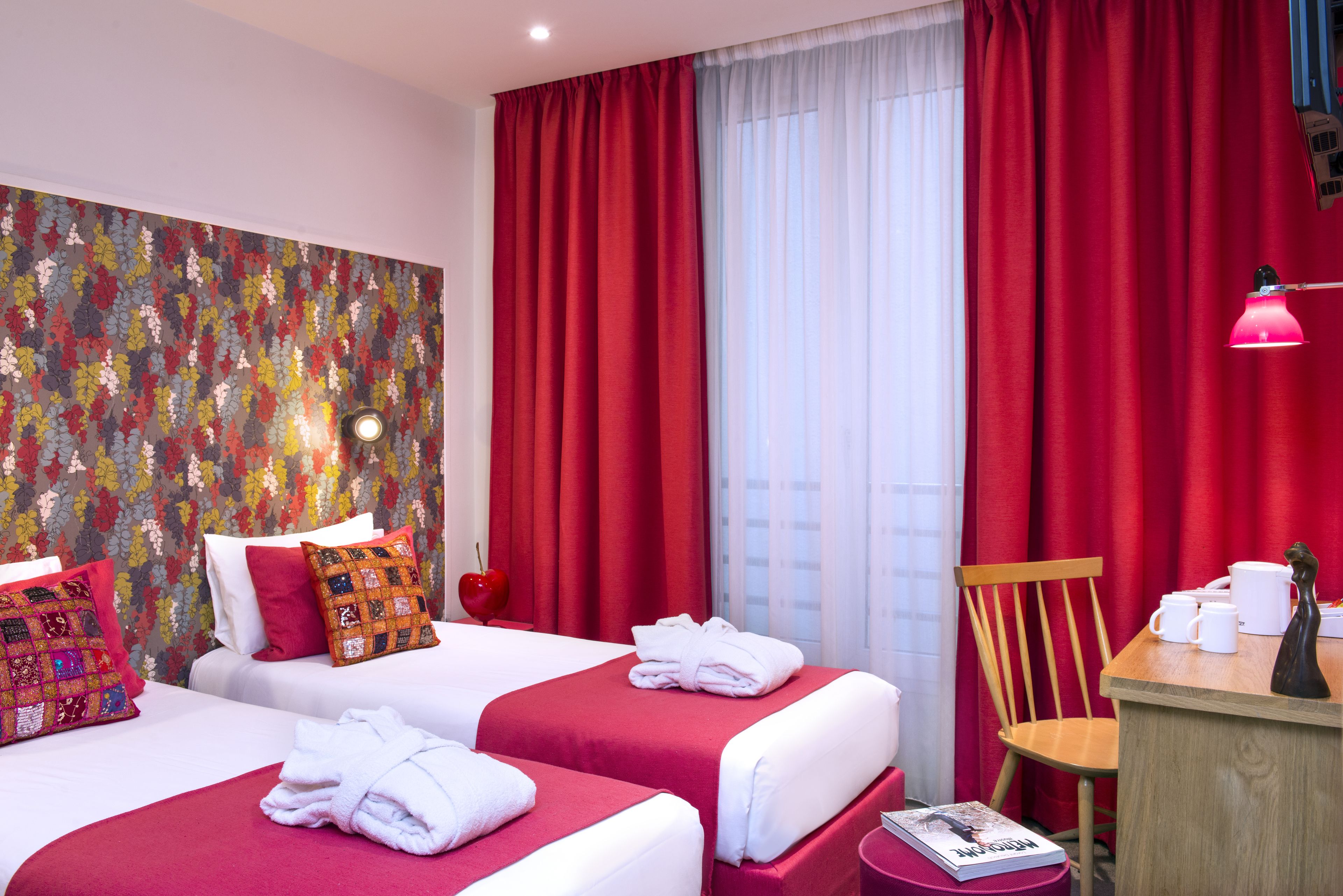 A hotel room with two beds, red curtains, a wooden chair, a small table with a lamp, and a floral wallpaper.