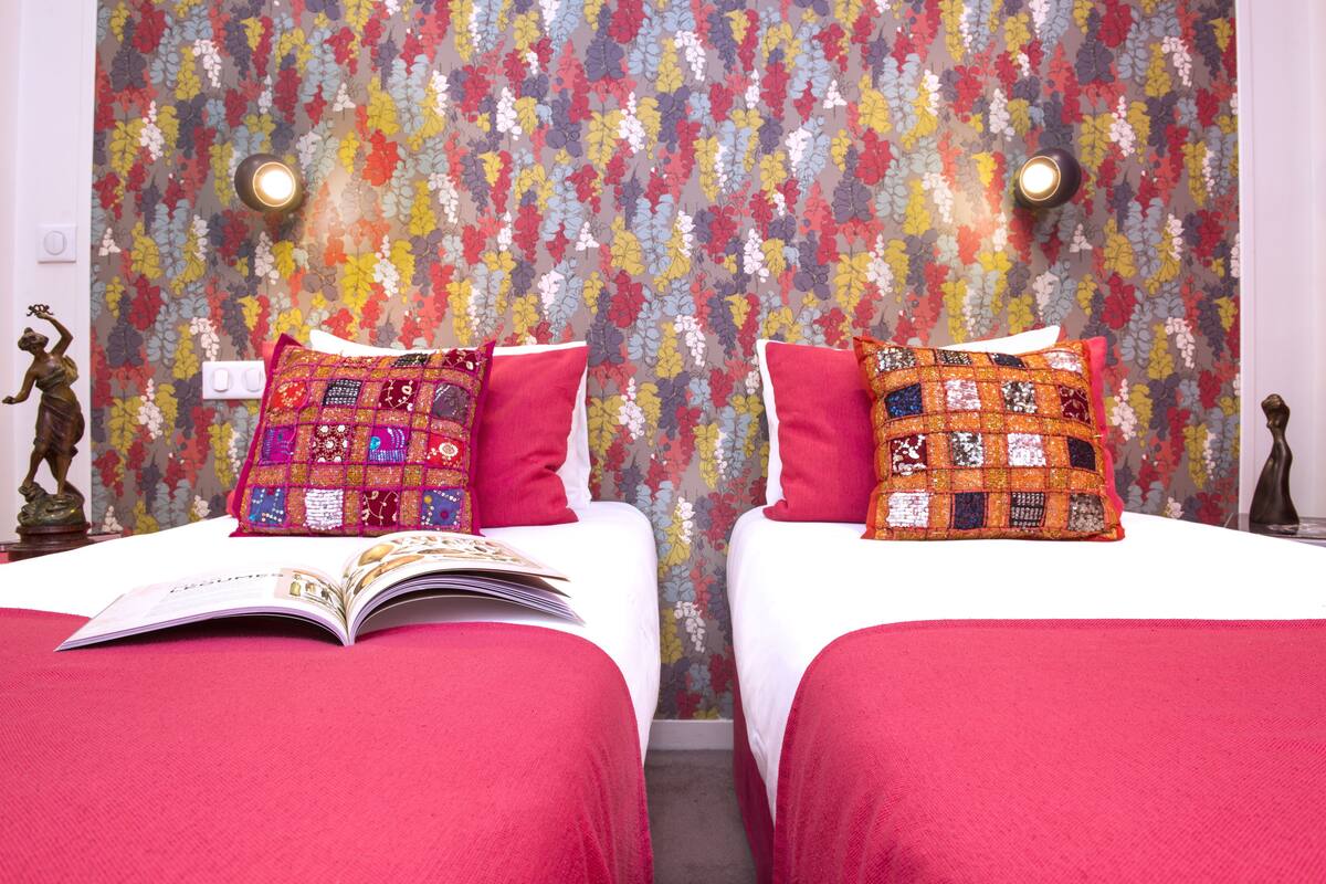 Hotellihuoneessa on kaksi sänkyä, joissa kummassakin on punainen päiväpeite ja tikattu tyyny. Huoneen seinillä on kukallinen tapetti ja siinä on kaksi seinään kiinnitettyä valaisinta.