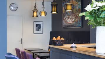 A restaurant interior with a wooden counter, purple and blue chairs, a basket of croissants, and a large white vase with flowers.