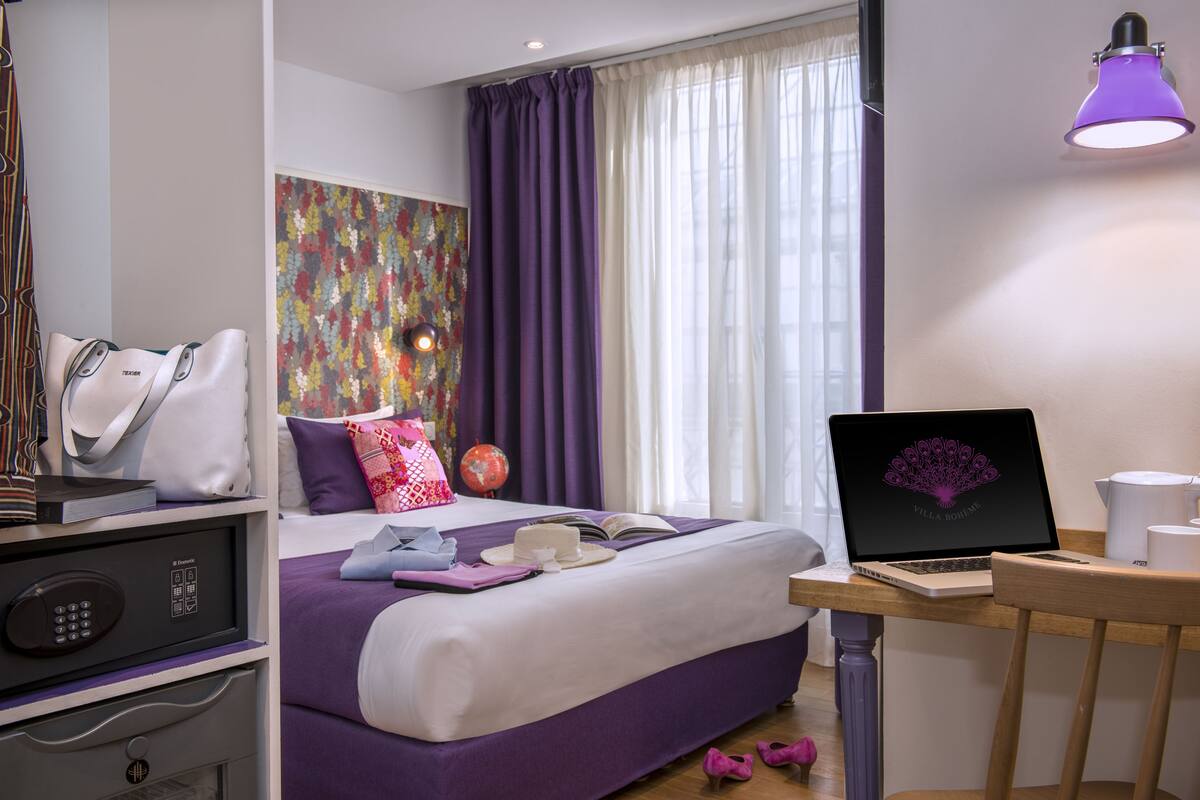 Moderni hotellihuone, jossa on sänky, työpöytä kannettavalla tietokoneella, maapallo ja matkalaukku.