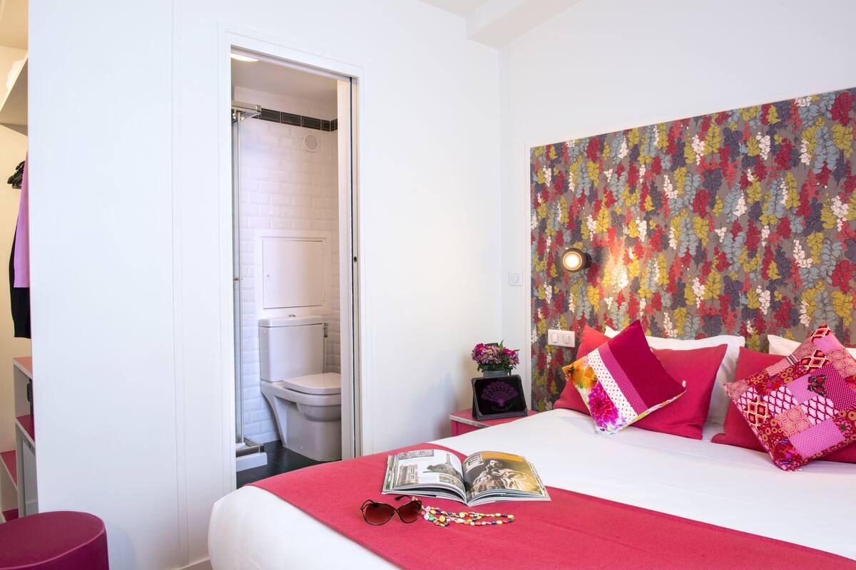 Hotellihuone, jossa on sänky, yöpöytä, punainen tuoli ja kylpyhuone, joka näkyy avoimesta ovesta.