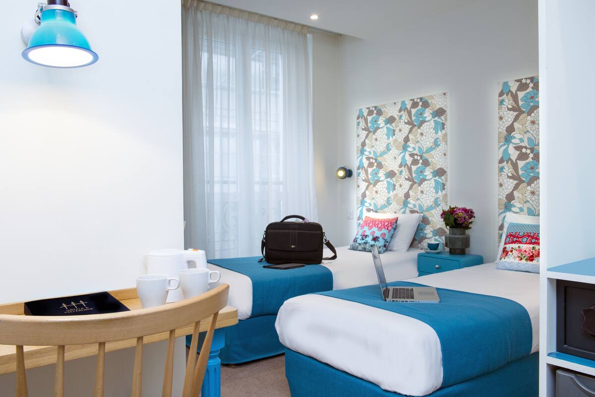 Moderni hotellihuone, jossa on kaksi sänkyä, työpöytä kannettavalla tietokoneella, tuoli, pöytä, jossa on kuppeja ja rasia, sekä suuri ikkuna, jossa on verhot.