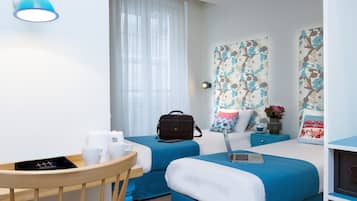 Moderni hotellihuone, jossa on kaksi sänkyä, työpöytä kannettavalla tietokoneella, tuoli, pöytä, jossa on kuppeja ja rasia, sekä suuri ikkuna, jossa on verhot.