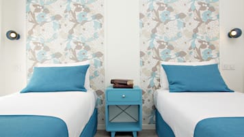 A room with two beds, each with blue bedding, a small bedside table with a book, and floral wallpaper.