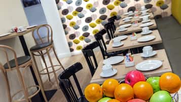 Un espace repas avec des tables dressées, des chaises et un panier de fruits contenant des oranges et des pommes.