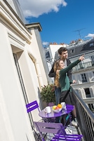 A balcony with a purple table and chairs, a potted plant, and a window overlooking a building.