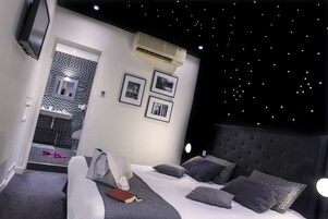A modern bedroom with a starry ceiling, a bed with grey bedding, and a small bedside table with a book and a lamp.