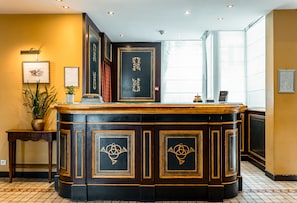 A reception area with a dark wooden counter, framed certificates on the wall, and a potted plant. - Villa Beaumarchais (Paris)