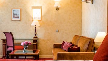 A hotel room with a bed, a wooden chair, a small table with a vase of flowers, a sofa with cushions, and two wall-mounted lamps.