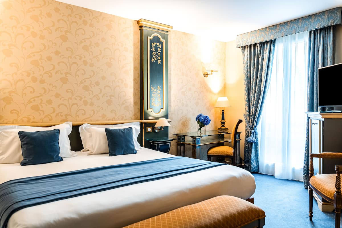 A hotel room with a large bed, a desk with a lamp, a chair, and a television.