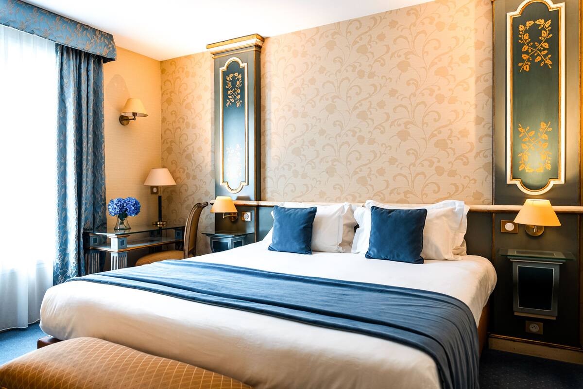 A hotel room with a large bed, two bedside tables with lamps, a nightstand, a mirror, and a wall with patterned wallpaper.