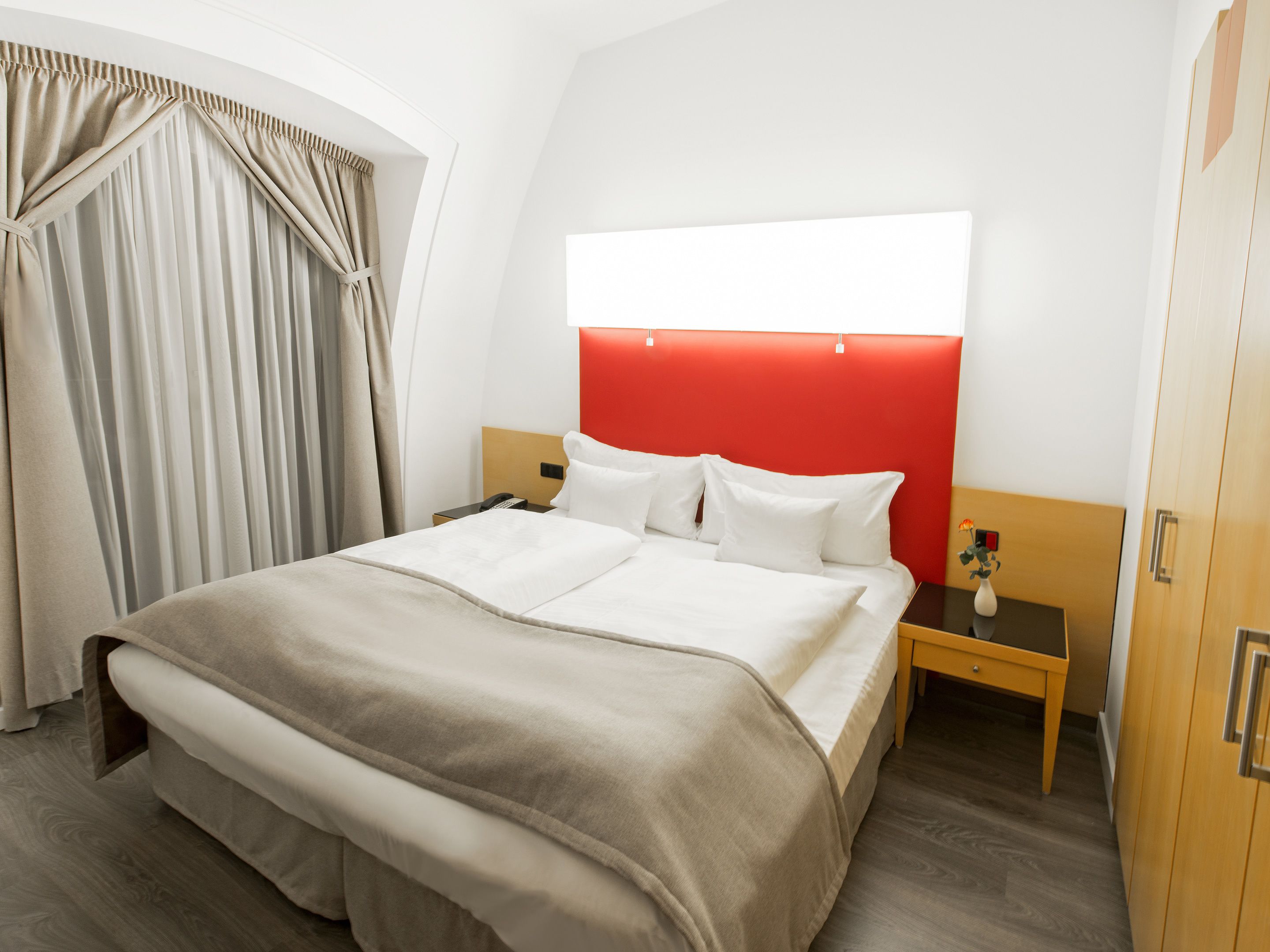 Habitació d'hotel amb un llit gran, una meseta de nit i una taula de nit amb un ram, i una paret vermella decorativa.