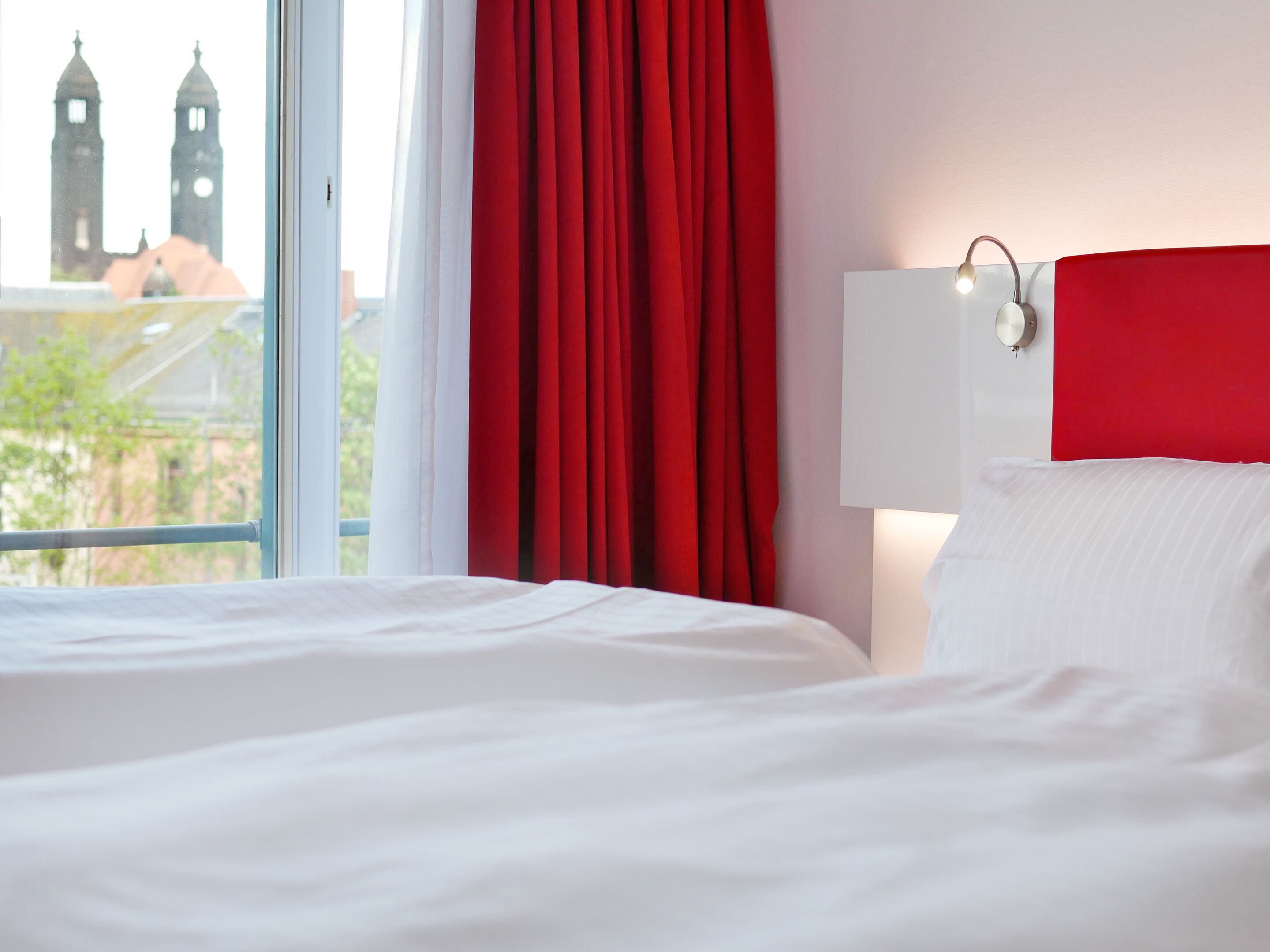 Ein Hotelzimmer mit einem großen Bett, einem Fenster mit roten Vorhängen und Blick auf zwei Kirchtürme.