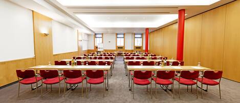 A conference room with rows of tables and red chairs, large windows, and a high ceiling.