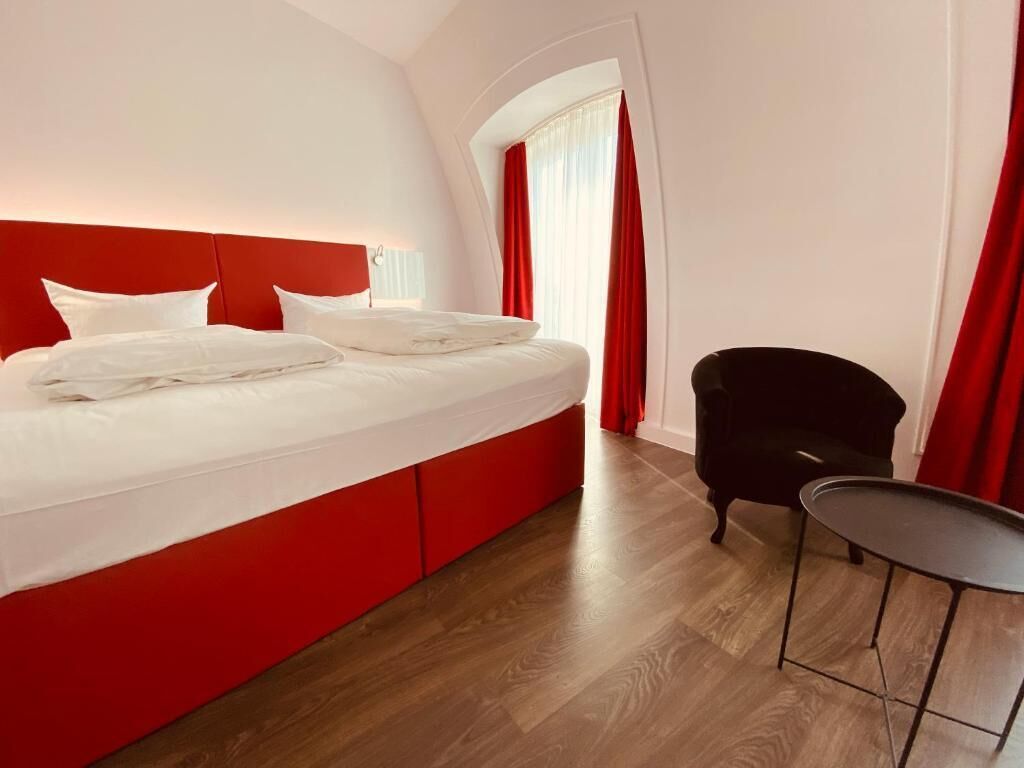 A hotel room with a large bed, a black armchair, a small table, and a window with red curtains.