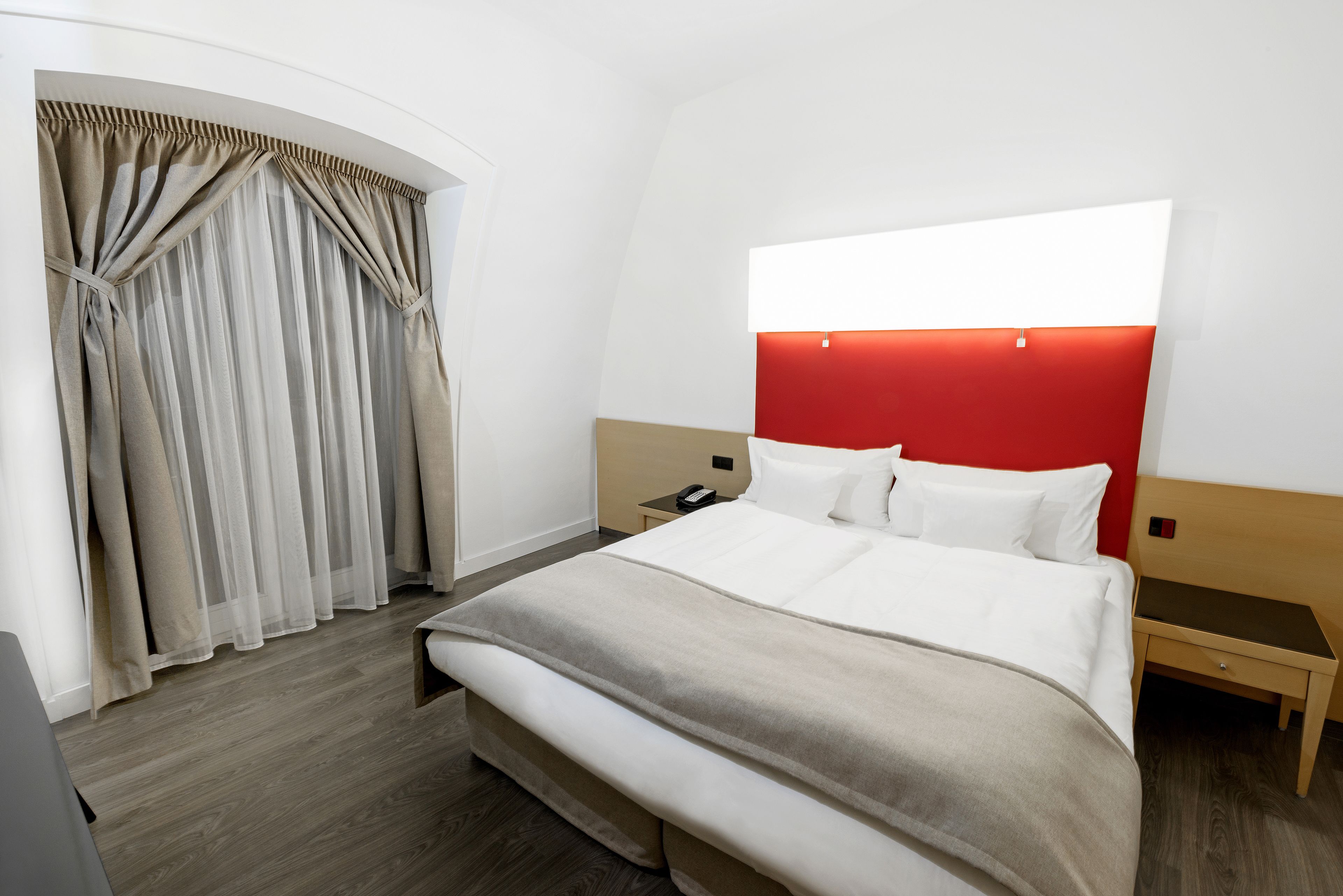 Una habitación de hotel con cama, mesita de noche, pared de acento roja y ventana con cortinas.
