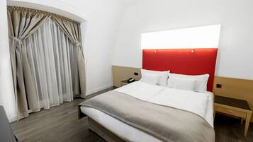 Una habitación de hotel con cama, mesita de noche, pared de acento roja y ventana con cortinas.