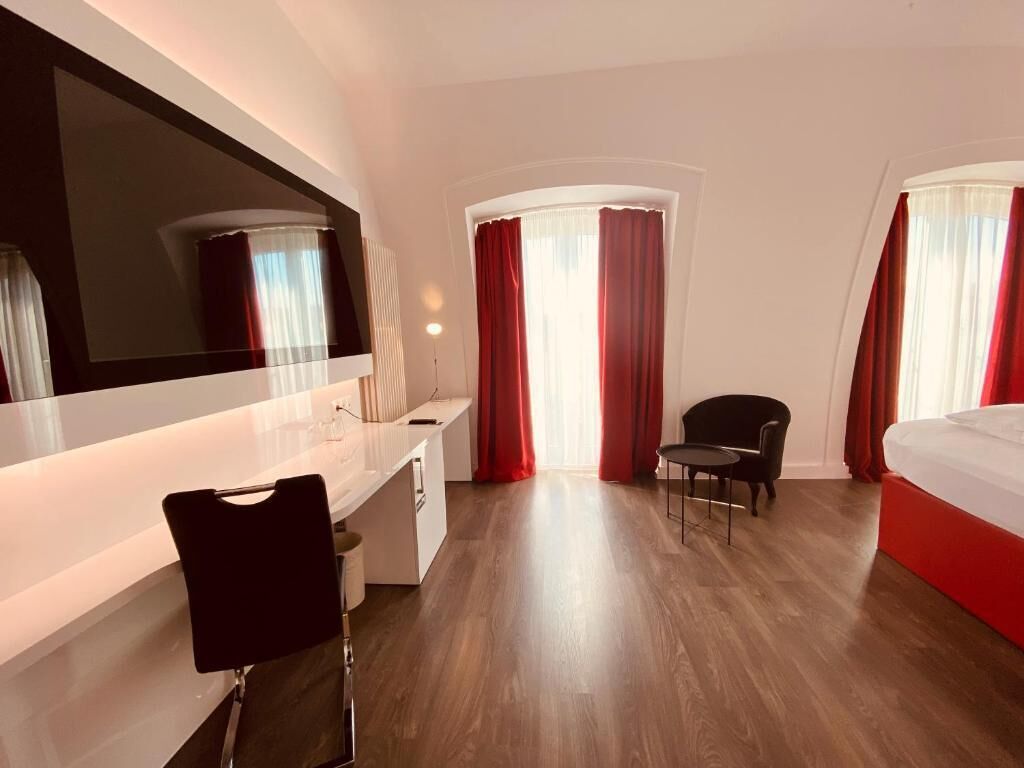 A modern hotel room with a large bed, a desk with a chair, a large mirror, and two windows with red curtains.