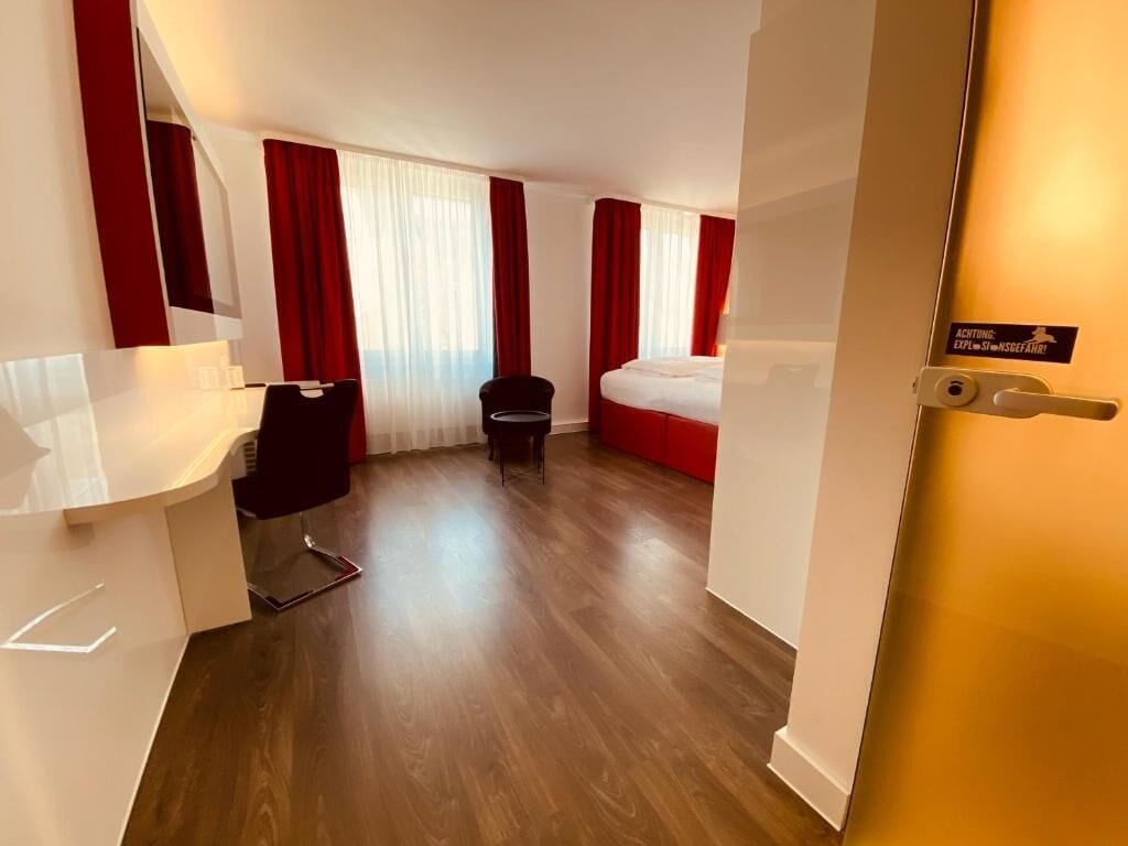 A hotel room with a bed, a desk, a chair, and a small table. There are curtains on the windows and a sign on the door.