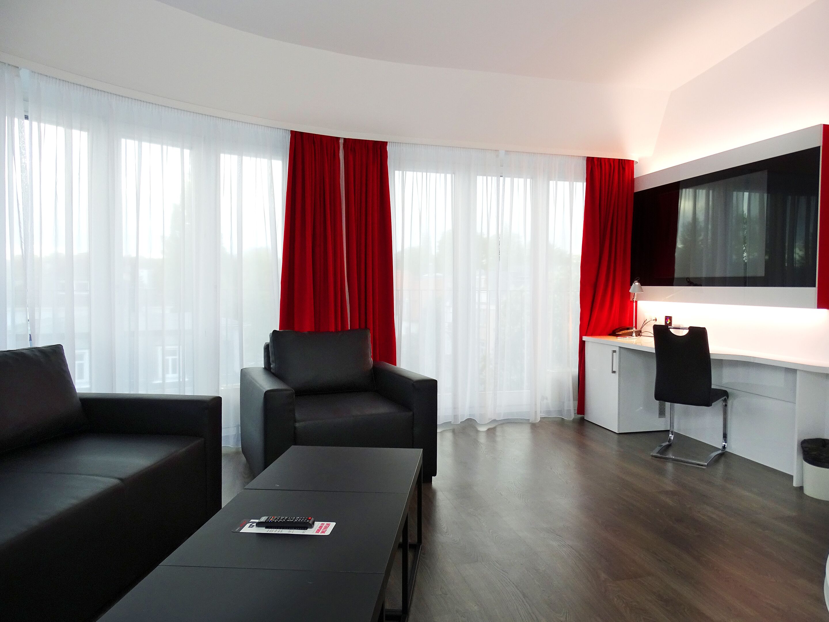 Een moderne hotelkamer met een donker houten vloer, een zwarte bank, een zwarte fauteuil, een klein tafeltje met een afstandsbediening en een bureau met een stoel.