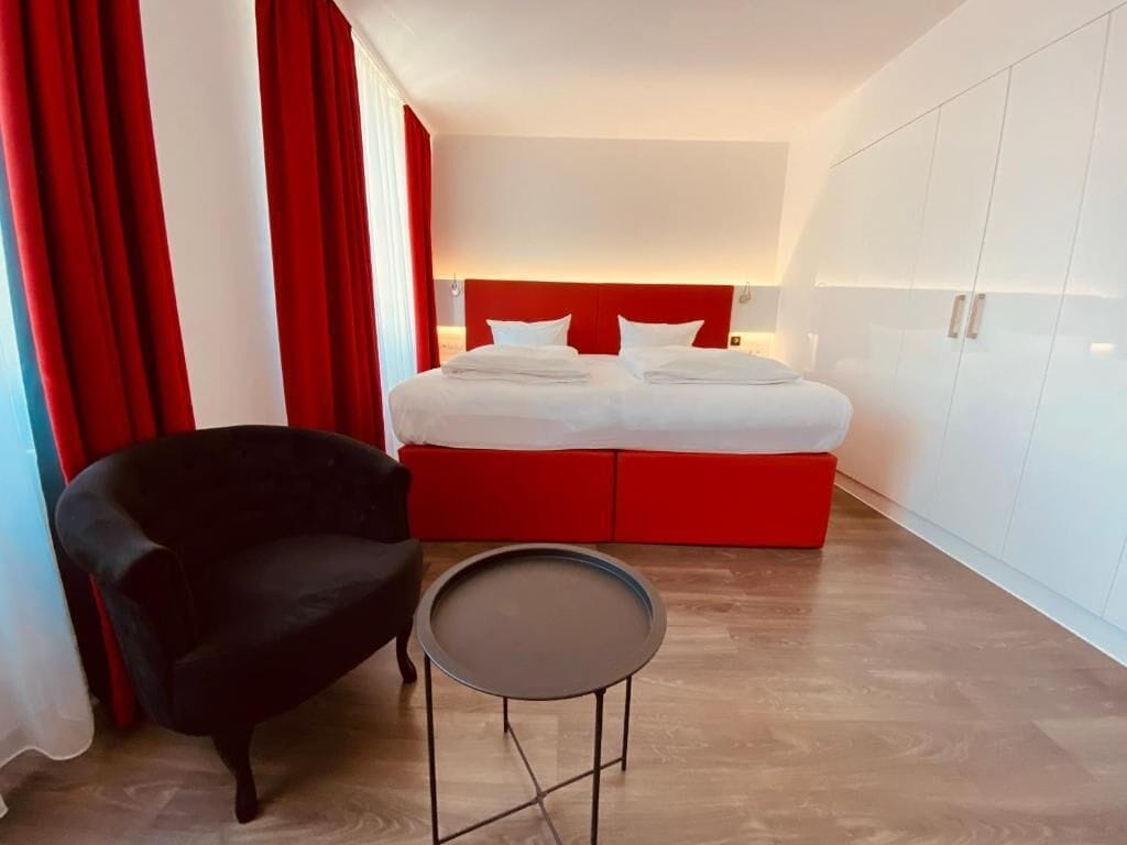 A modern hotel room with a large bed, red accents, a black armchair, and a small table.
