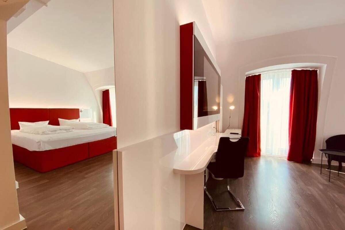 Una habitación de hotel moderna con una cama roja, un escritorio blanco, un espejo y vistas al exterior.