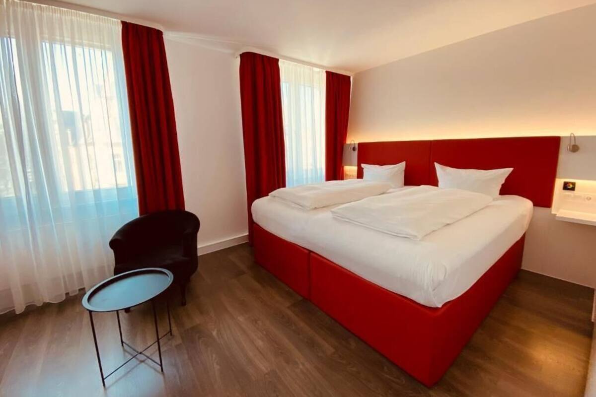 Una habitación de hotel con una cama roja, una mesita pequeña, una silla y una ventana con cortinas.