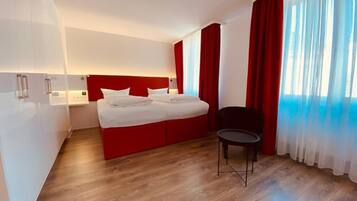 A modern hotel room with a red accent wall, a large bed, a small table, and a view of the city through sheer curtains.
