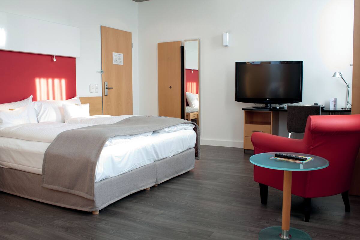 Una habitación de hotel con una cama, un televisor, un sillón rojo y una mesita.