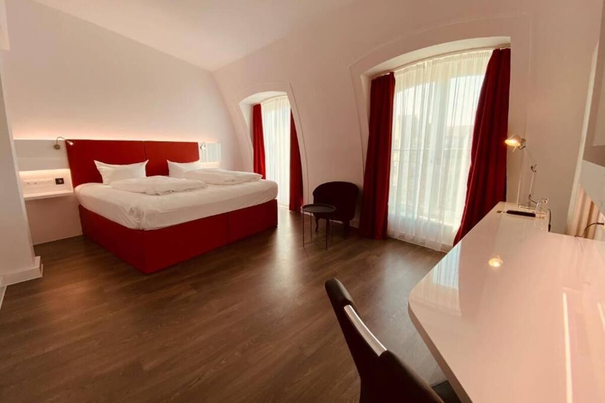 Habitación de hotel moderna con una cama de detalles rojos, una zona de escritorio separada y un ventanal con cortinas.