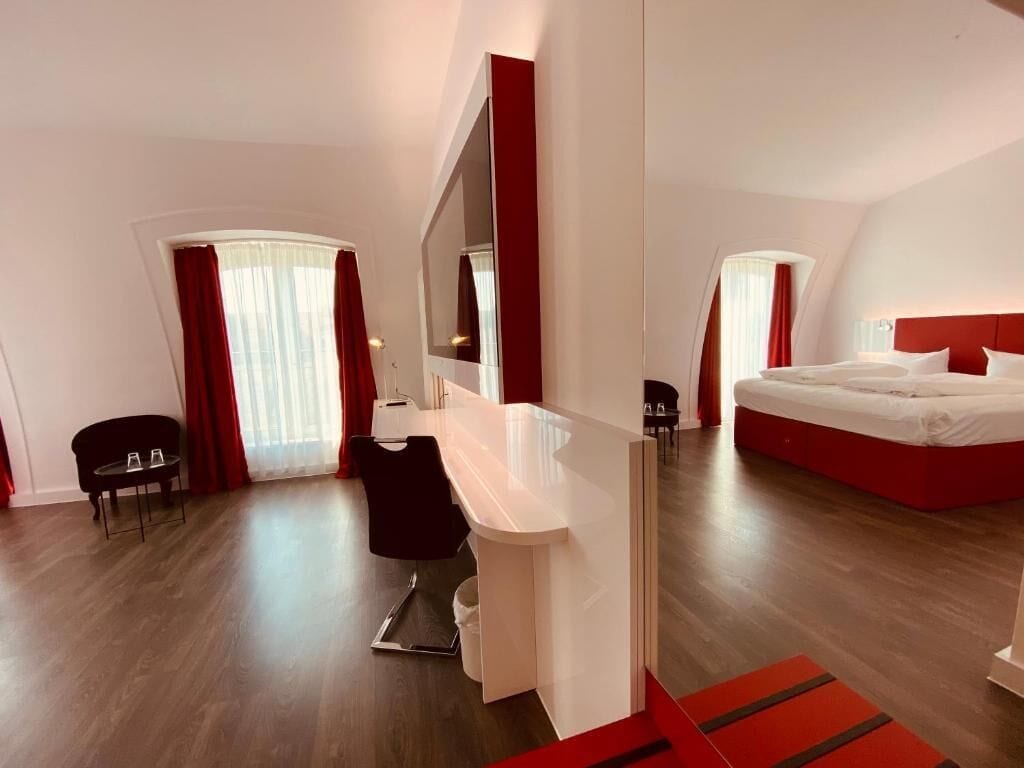 Una camera d'albergo moderna con un letto grande, una scrivania e una sedia. È presente un tavolino con due bicchieri e un'altra sedia. La stanza ha uno specchio grande e una parete d'accento rossa.