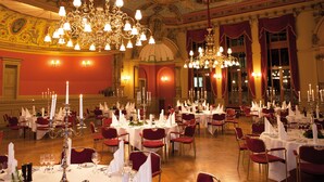 Een statige balzaal met gedetailleerde kroonluchters, rode bekleden stoelen en gedekte tafels voor een formeel evenement.