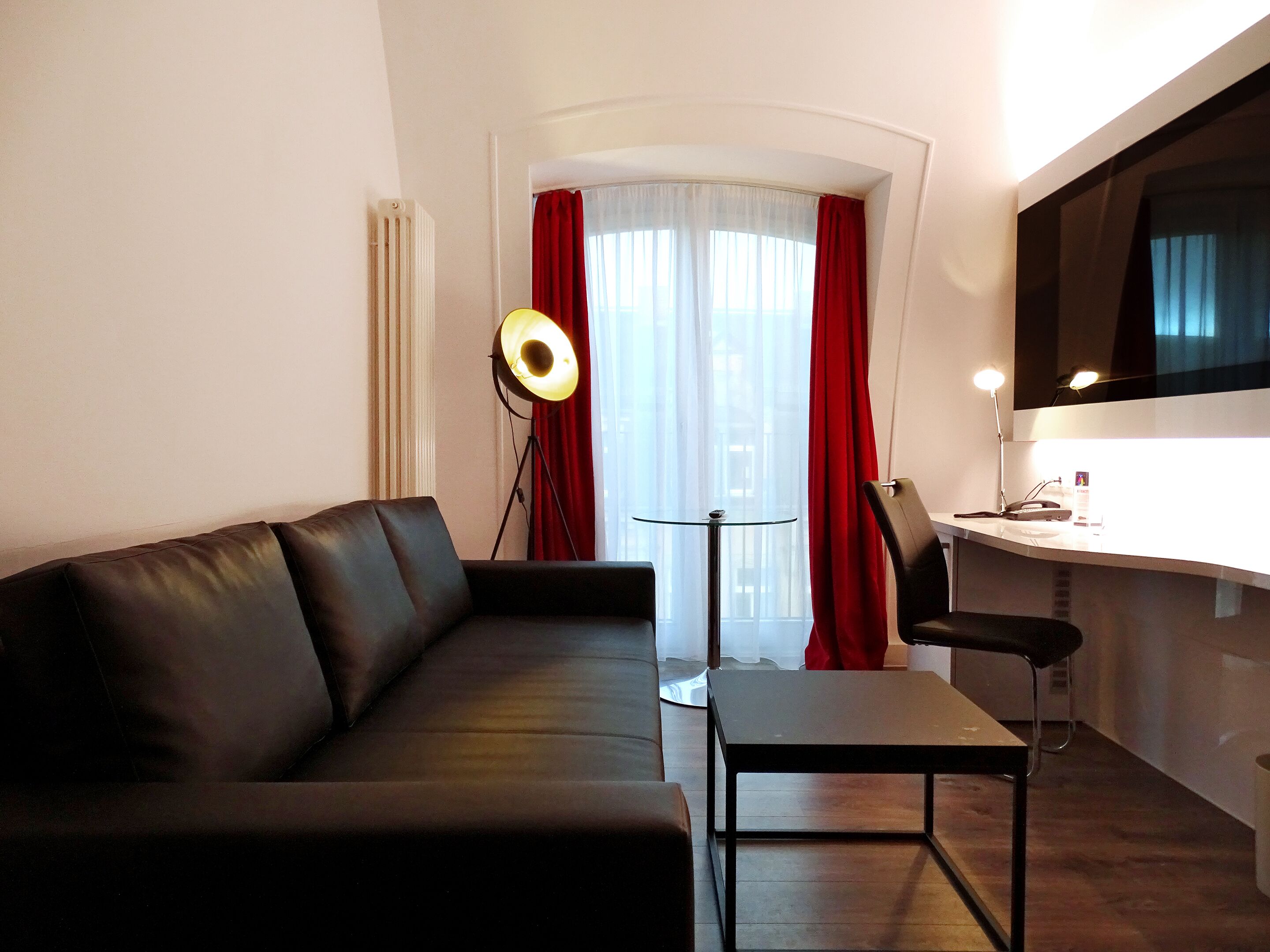 A modern living room with a black leather sofa, a glass coffee table, a desk with a chair, and a large window with red curtains.