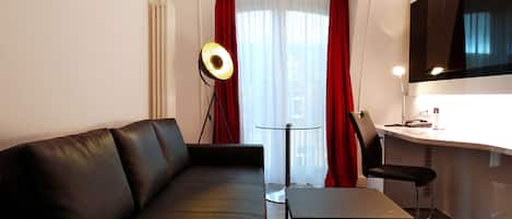 A modern living room with a black leather sofa, a glass coffee table, a desk with a chair, and a large window with red curtains.