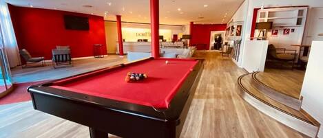 A pool table in a modern room with a red accent wall, a seating area, and a staircase leading to an upper level.