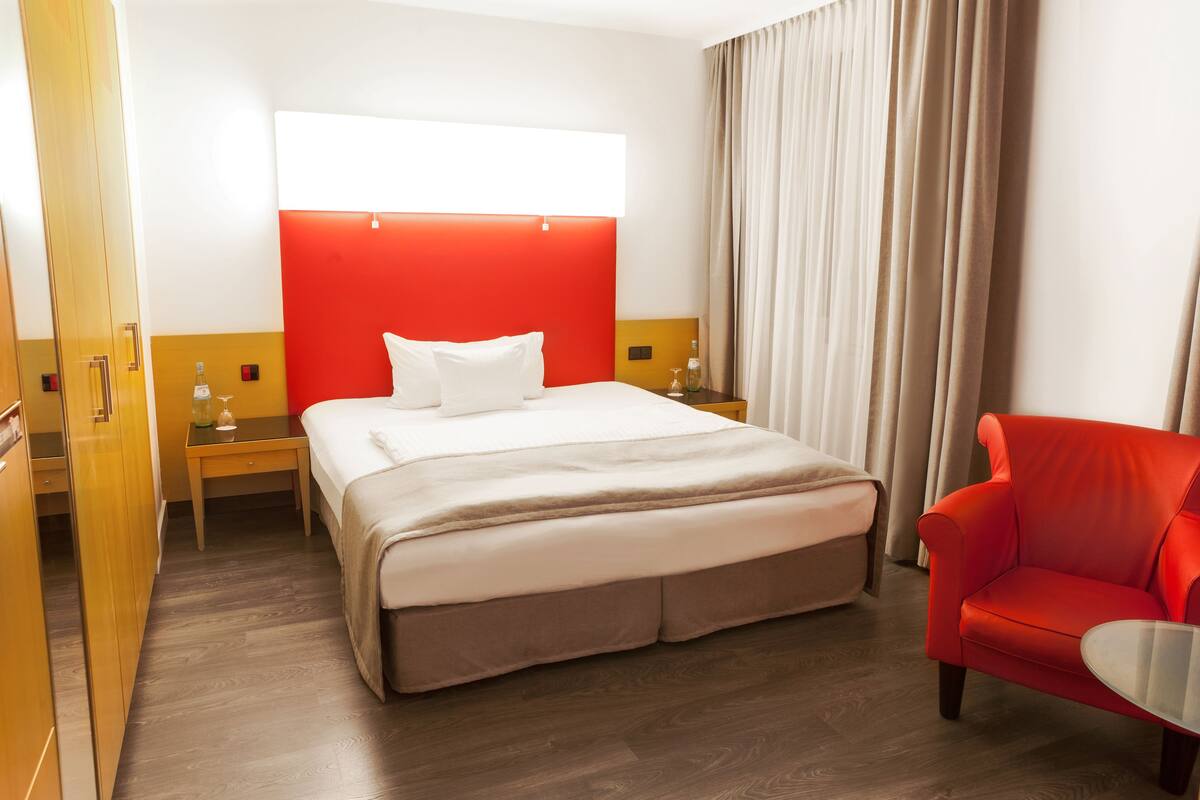 Una habitación de hotel con una cama, un sillón rojo, una mesita de noche y un armario de madera.