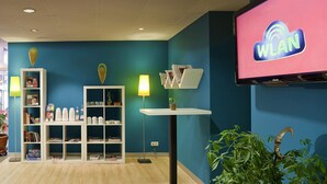 A room with a blue wall, a white bookshelf, a table with a lamp, a TV screen displaying 'WLAN', and a plant. - ibis Styles Düsseldorf - Neuss (Neuss)