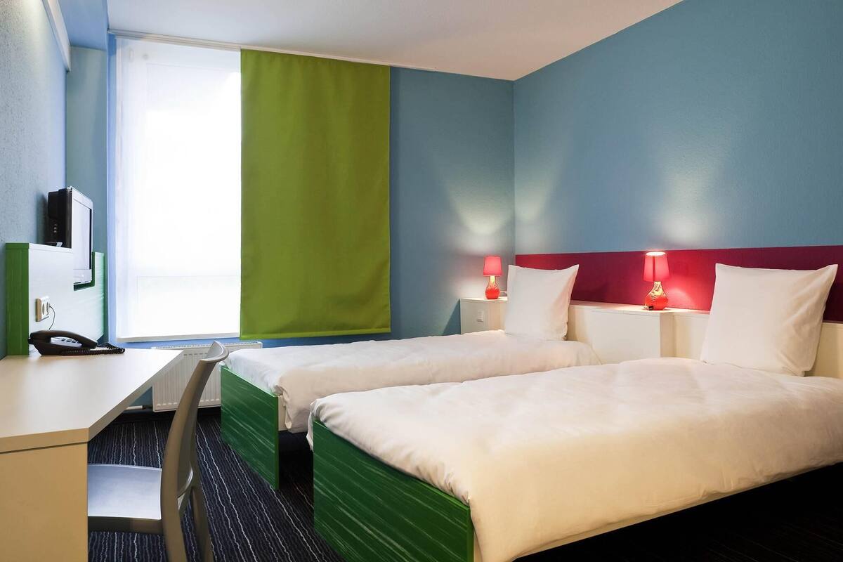 A hotel room with two beds, a desk, a chair, and a window with curtains.