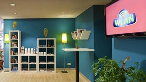 A room with a blue wall, a white bookshelf, a table with a lamp, a TV screen displaying 'WLAN', and a plant. - ibis Styles Düsseldorf - Neuss (Neuss)