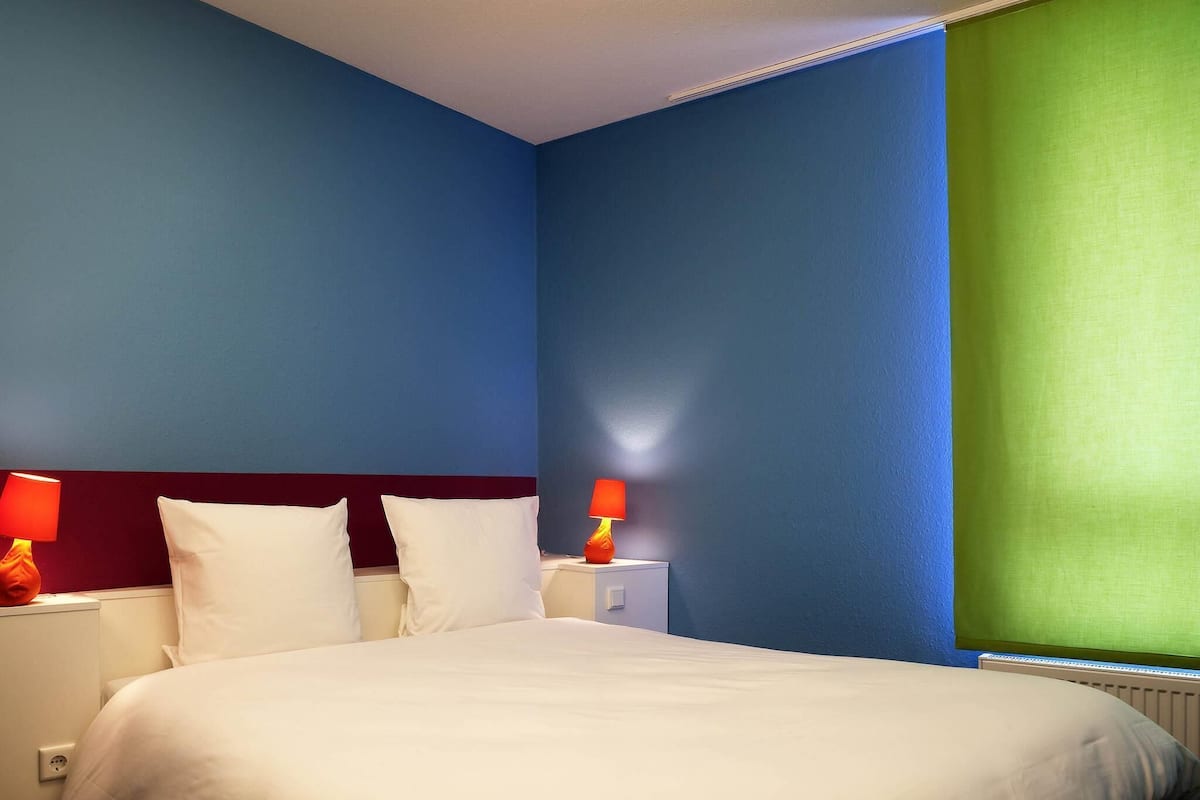 A hotel room with a bed, two bedside tables with lamps, and a window with a green blind.