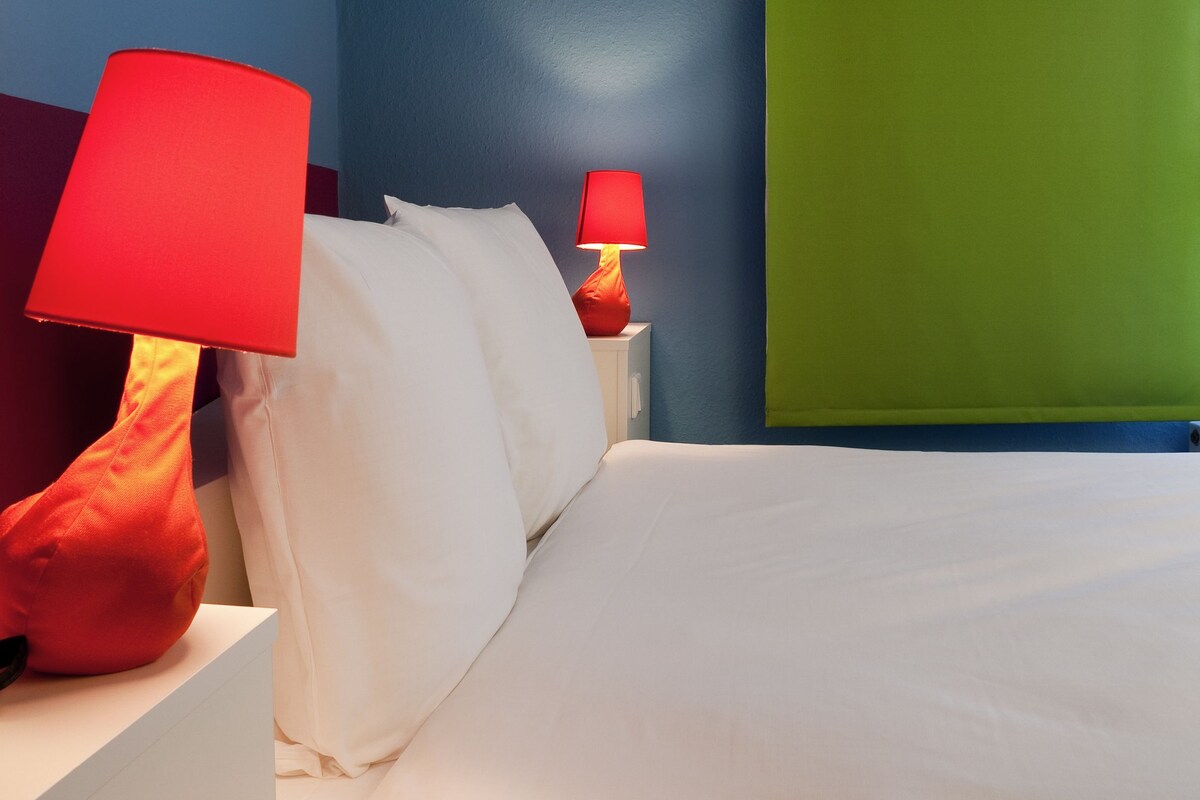 A neatly made bed with white linens, two bedside tables with red lamps, and a blue and green wall.