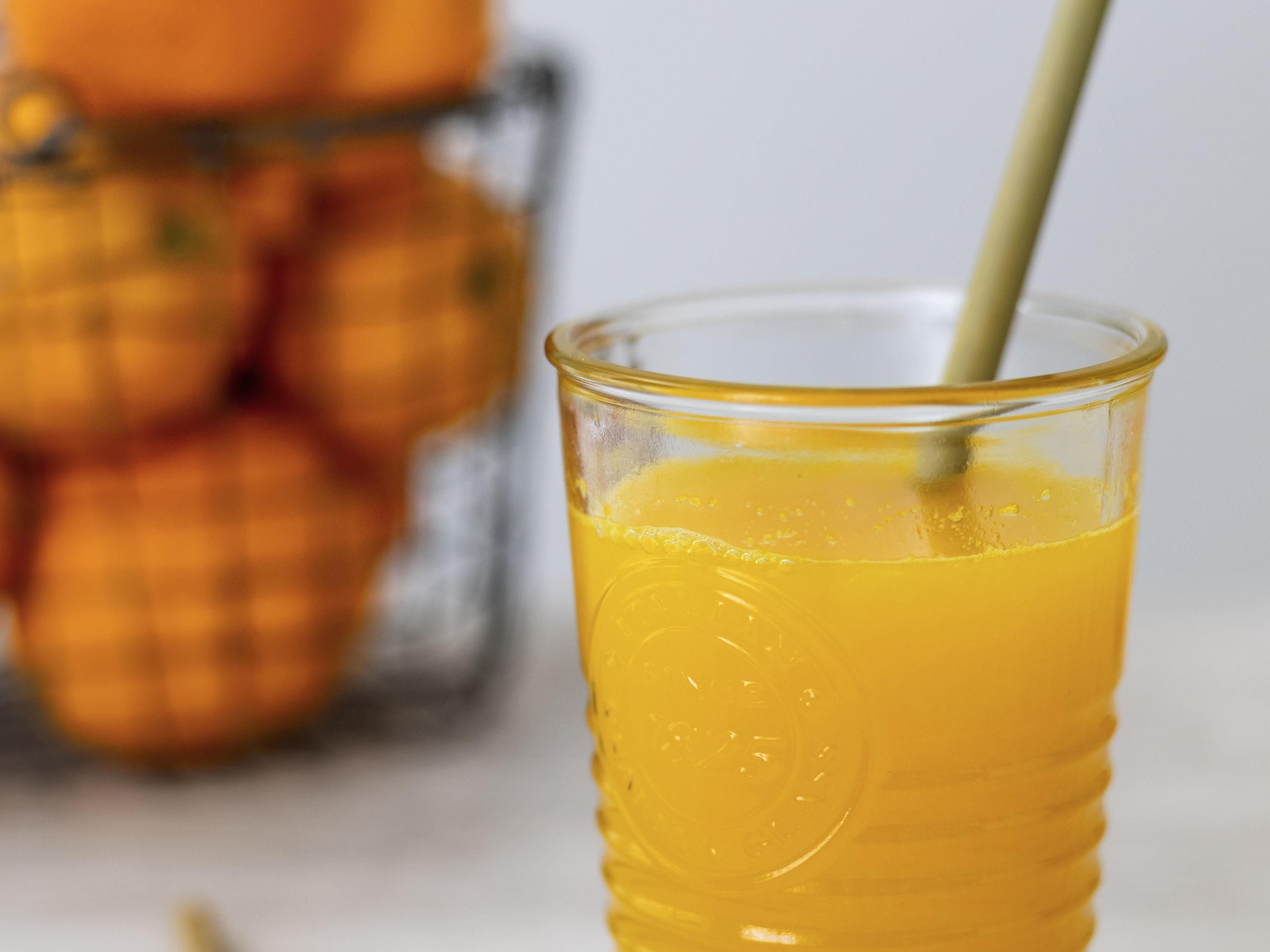 Un vaso de jugo de naranja con una pajita, y un fondo desenfocado de naranjas.