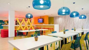 Un comedor luminoso con mesas blancas y sillas de colores, una pantalla de televisión y un papel tapiz estampado en el fondo.