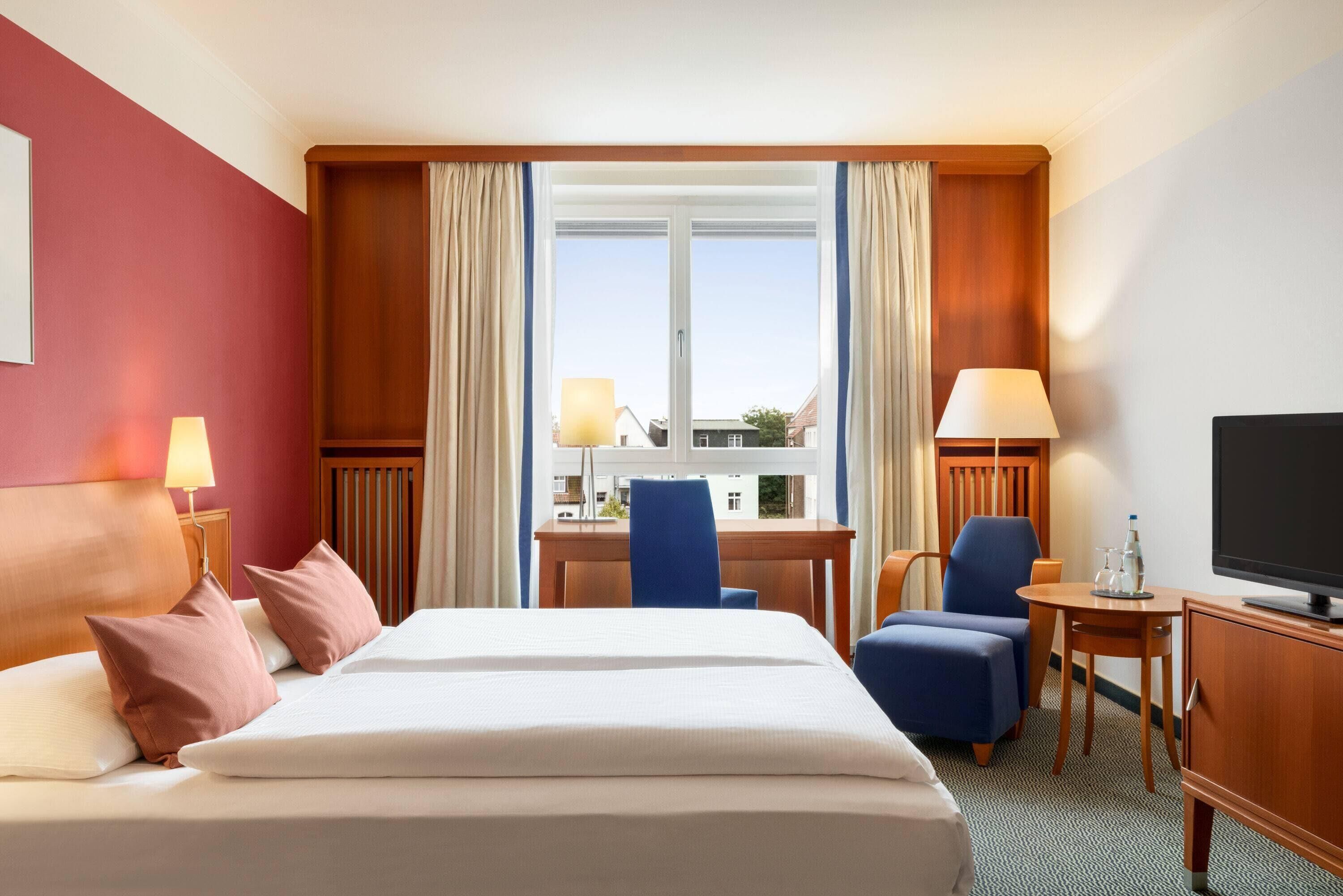 Hotellihuone, jossa on sänky, kaksi tuolia, työpöytä, televisio ja ikkuna, jossa on verhot.