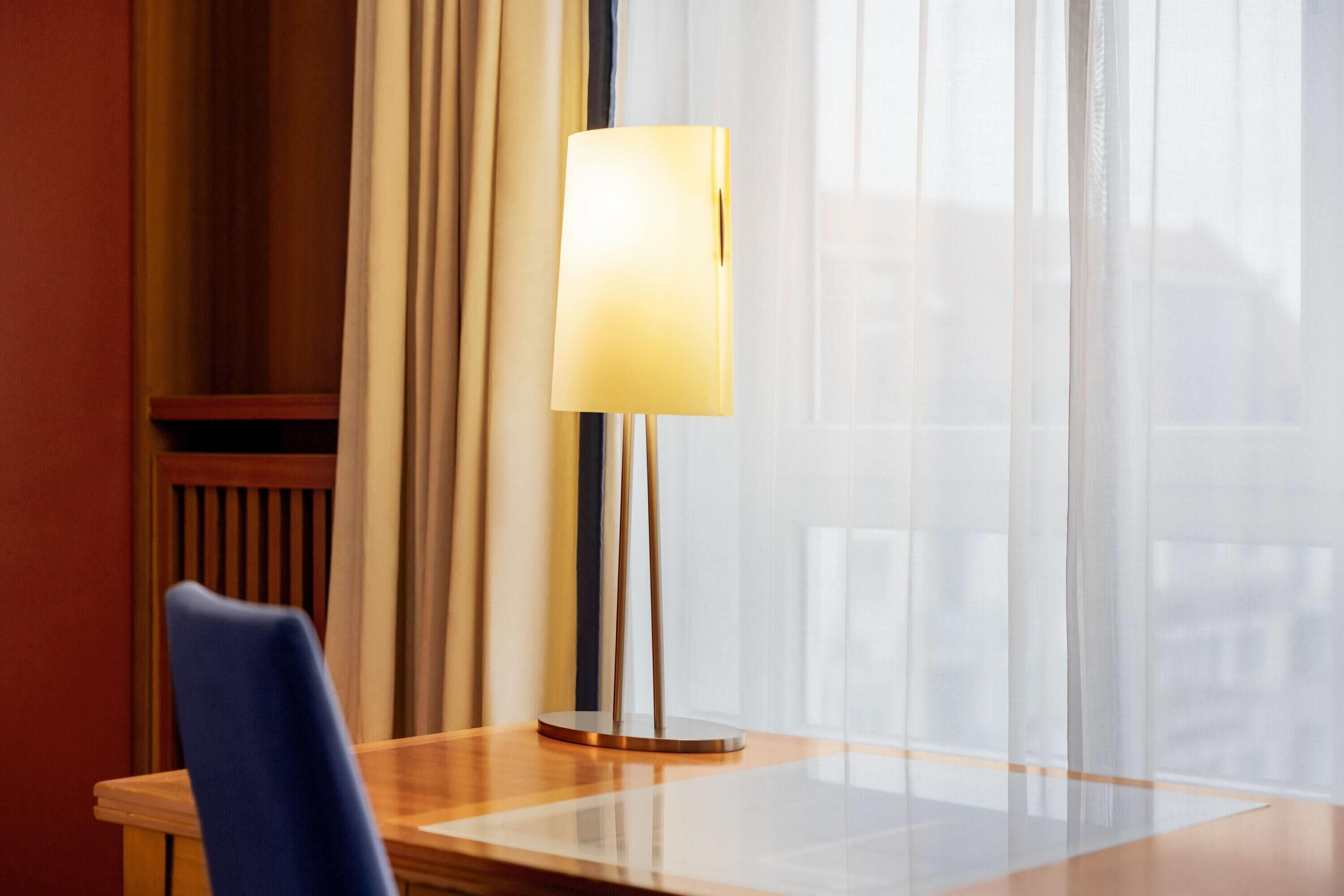 A hotel room with a table, a chair, a lamp, and a window with sheer curtains.